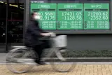 En kvinna med skyddsmask cyklar förbi en monitor med aktiekurser i Tokyo