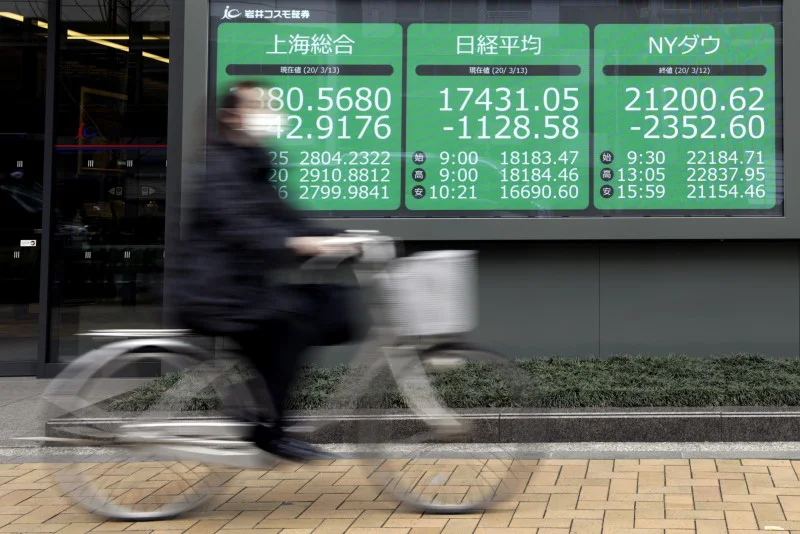 En kvinna med skyddsmask cyklar förbi en monitor med aktiekurser i Tokyo